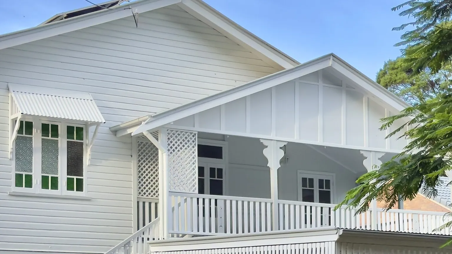 A white timber Queenslander house with a lattice verandah and green-tinted sash windows behind a white picket fence in Sherwood, Brisbane