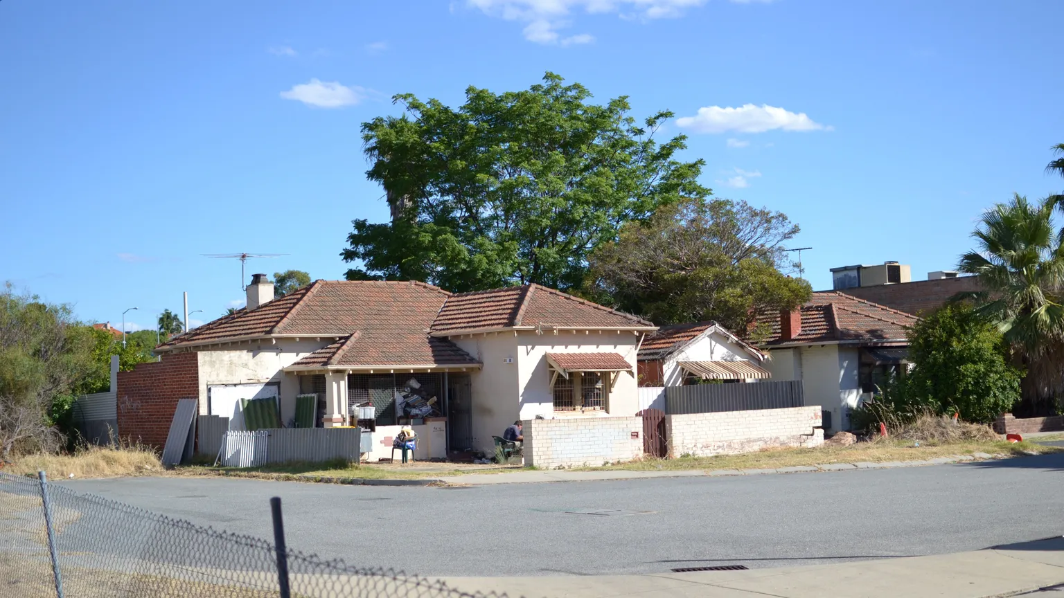 Wide view of an older single-storey brick and tile suburban house on a corner block beside a sealed road