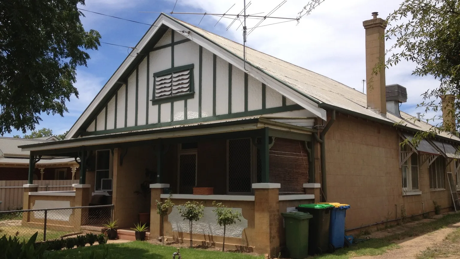 An older rendered dwelling with a steep gabled roof, chimney, deep front veranda and low masonry front fence on a New South Wales residential street