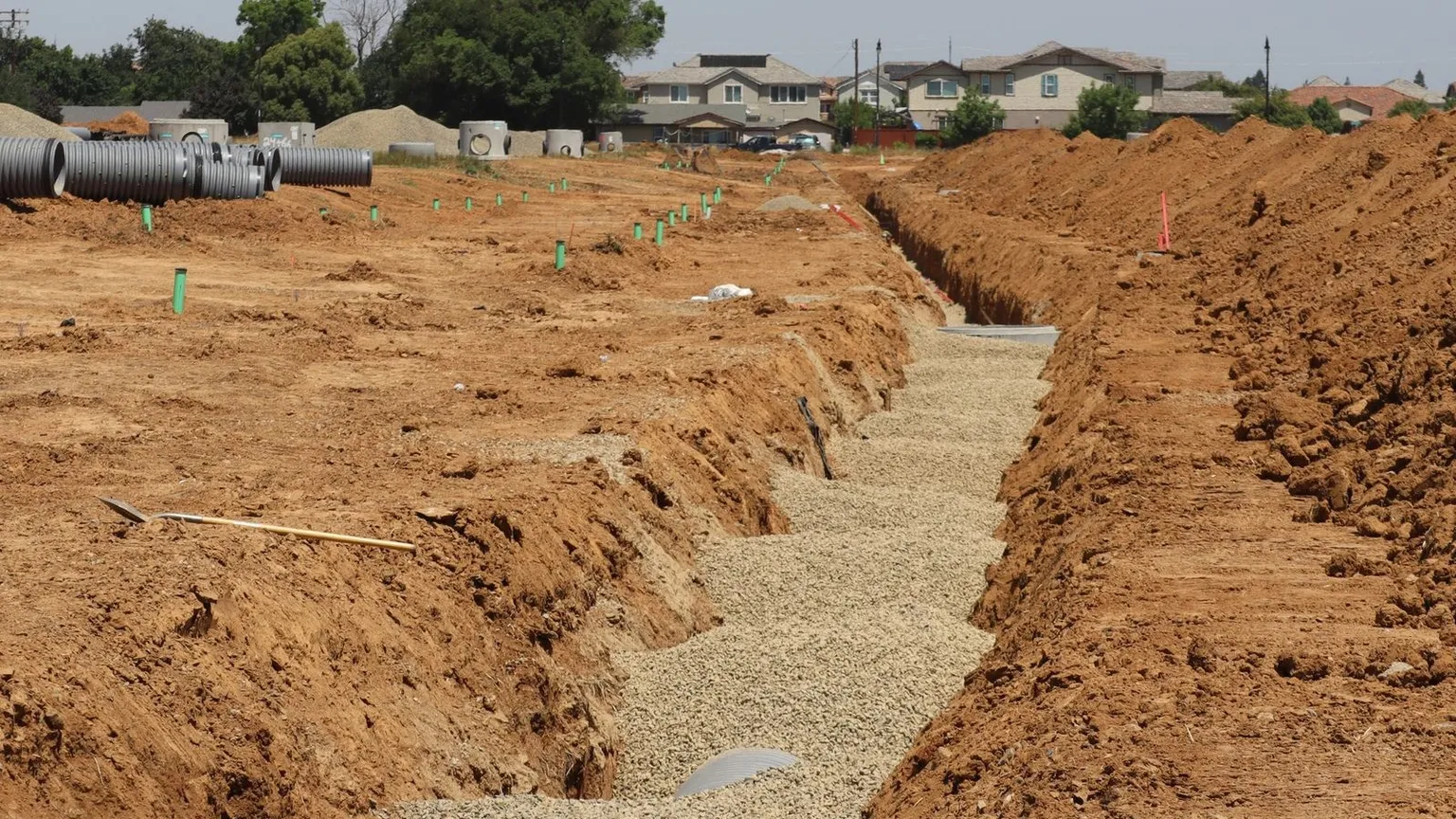 Subdivision under construction with a deep services trench, gravel bedding and pipes, and graded housing lots beyond