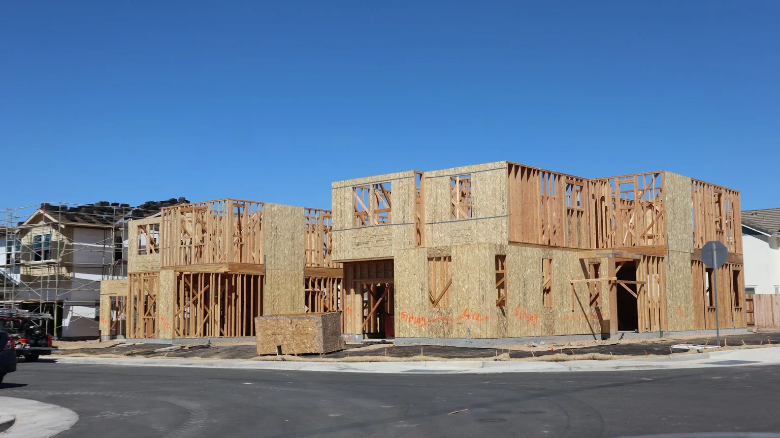 Wide view of several new detached houses at timber frame stage on a suburban street under a clear blue sky