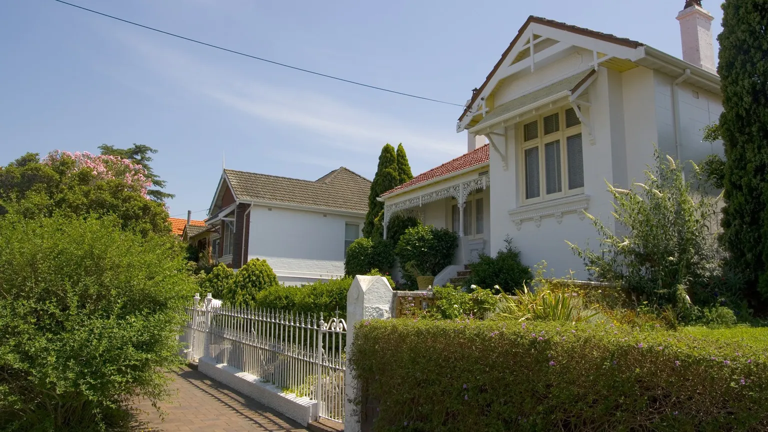 A quiet suburban street with a white rendered house behind a low picket fence and green hedges, seen on a clear day