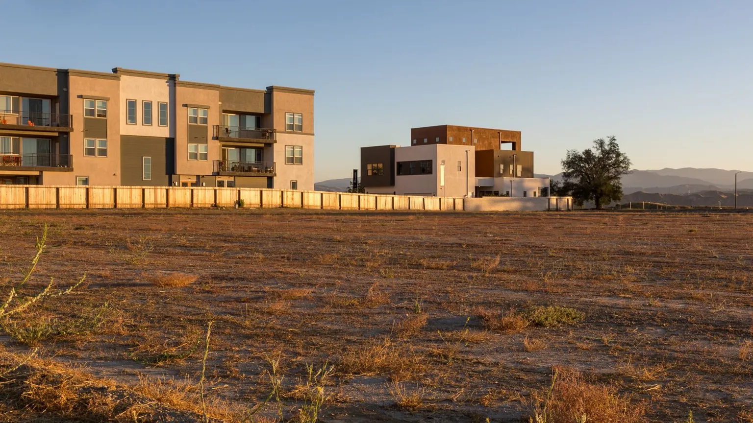 Bare undeveloped land parcel with dry grass in front of new townhouses and a boundary fence