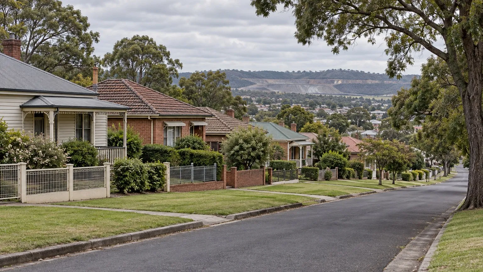 Older single-storey weatherboard and brick cottages with low front fences on gently sloping ground along a quiet suburban street, with sealed road, concrete kerb, grass verge and mature street trees under soft overcast light