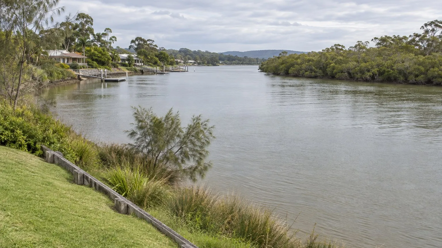 Wide tidal river curving away between low grassy banks with casuarina and native grass along the water's edge, mangroves on the far shore and single-storey houses beyond the tree line under an overcast sky
