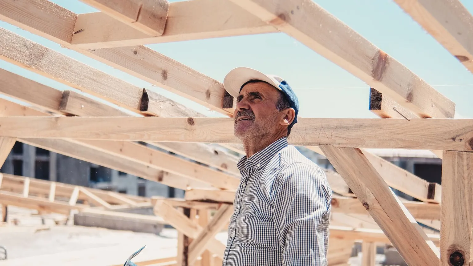 Person wearing a cap and checked shirt looking up at timber roof framing on a house under construction