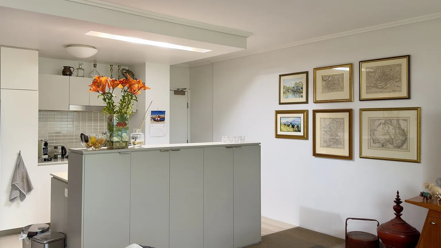 A light-filled apartment kitchen with a central island benchtop, tiled splashback, wall oven and overhead cabinetry.