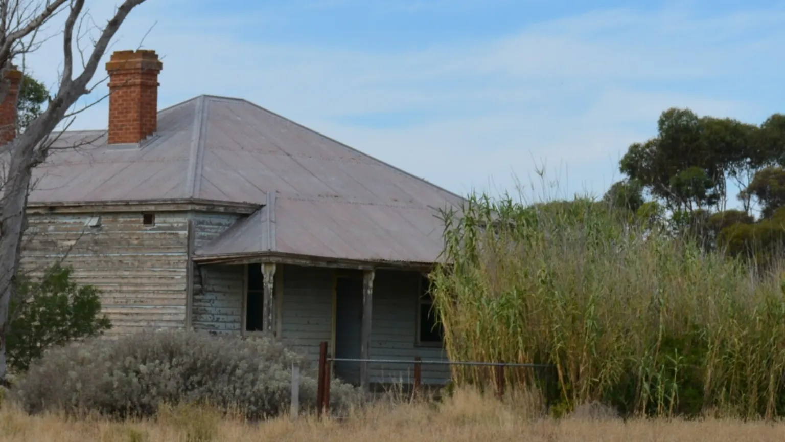 Weathered weatherboard cottage with a corrugated iron roof and brick chimney on an unfenced country block, with dry grass and scrub around it
