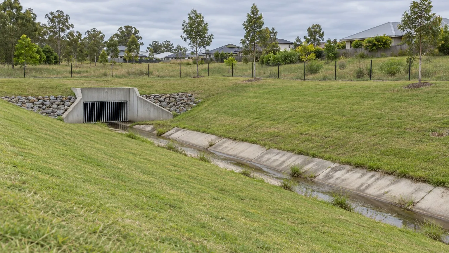 Broad grassed suburban stormwater drainage basin with a concrete channel, headwall and grate and a low wire fence on an overcast day
