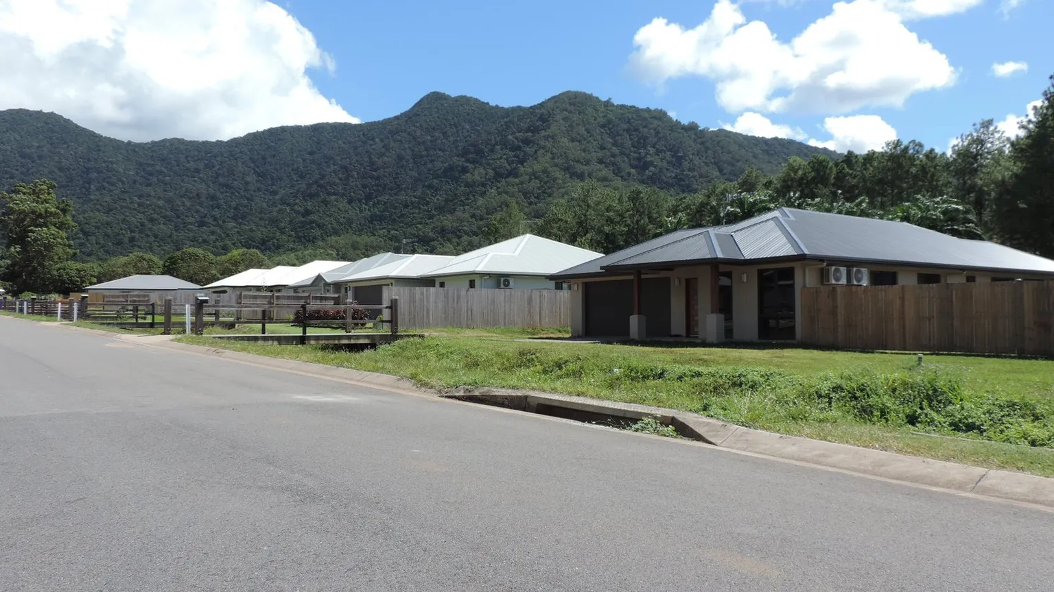 A residential street of completed new low-set houses with metal roofs, carports and timber fences in Edmonton, Queensland, under a mountain backdrop
