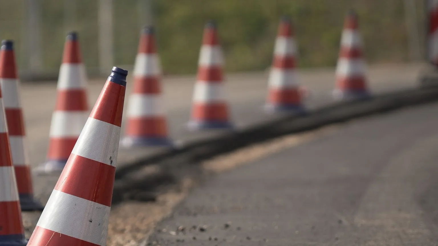 A receding line of red and white traffic cones closing off the edge of a sealed asphalt road beside a low barrier