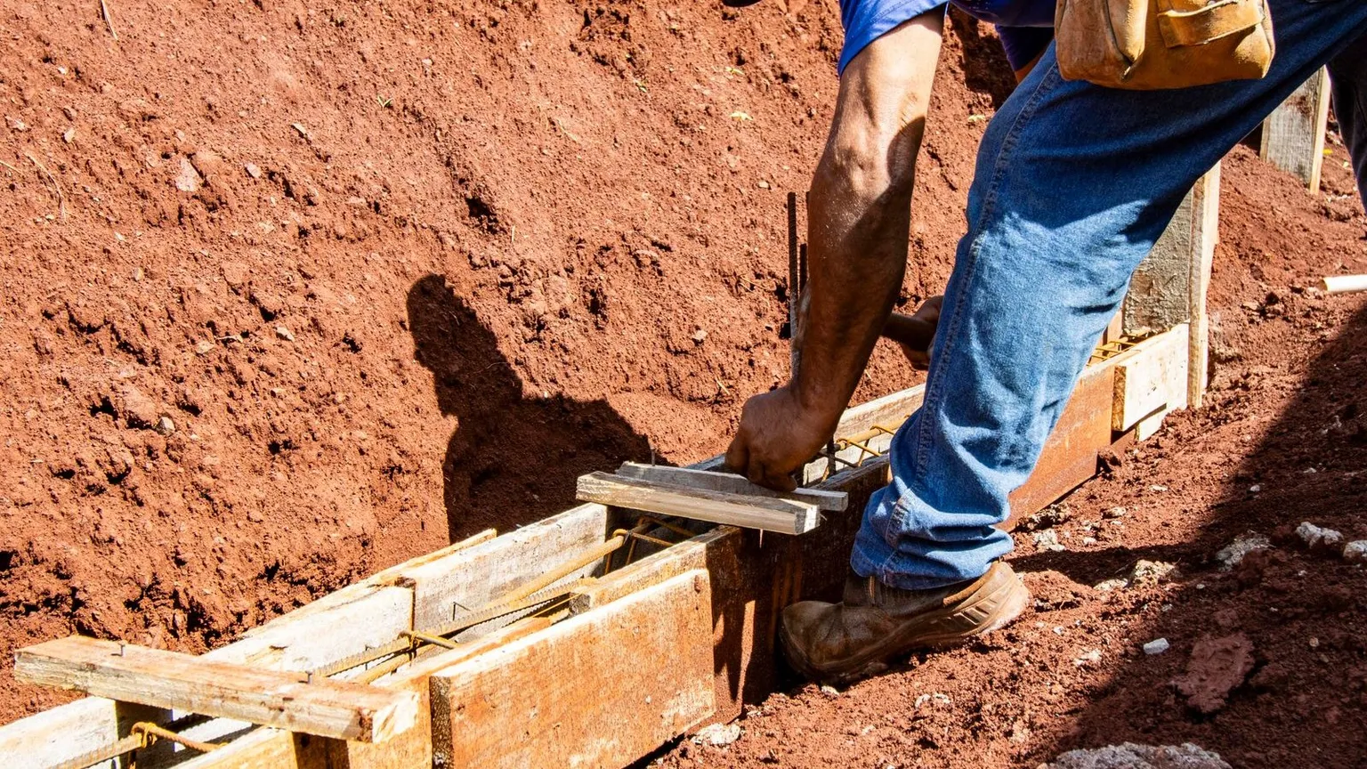 A worker in work boots and jeans setting timber formwork and steel reinforcement for a strip footing in red-brown clay soil