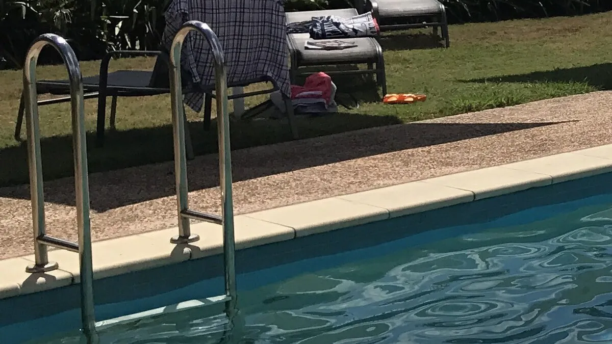 A residential swimming pool with clear blue water beside a tiled pool edge, a stainless steel entry ladder, mown lawn and outdoor lounge seating beyond, in daylight