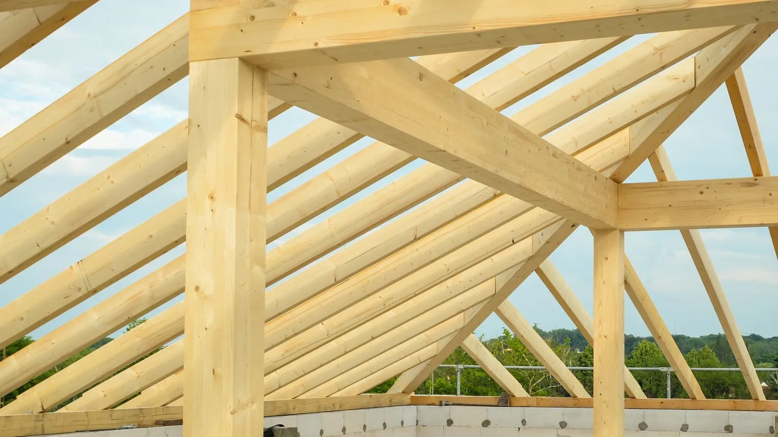 A newly erected timber roof frame and top plates under construction above a concrete slab edge, with sky visible through the trusses